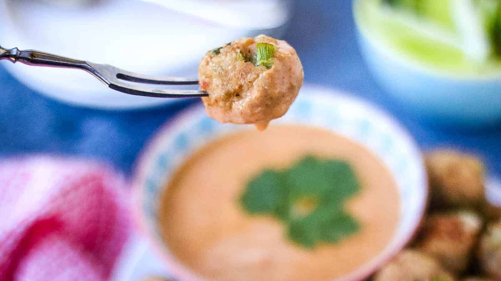A fork holds a meatball above a bowl of creamy dipping sauce garnished with cilantro.