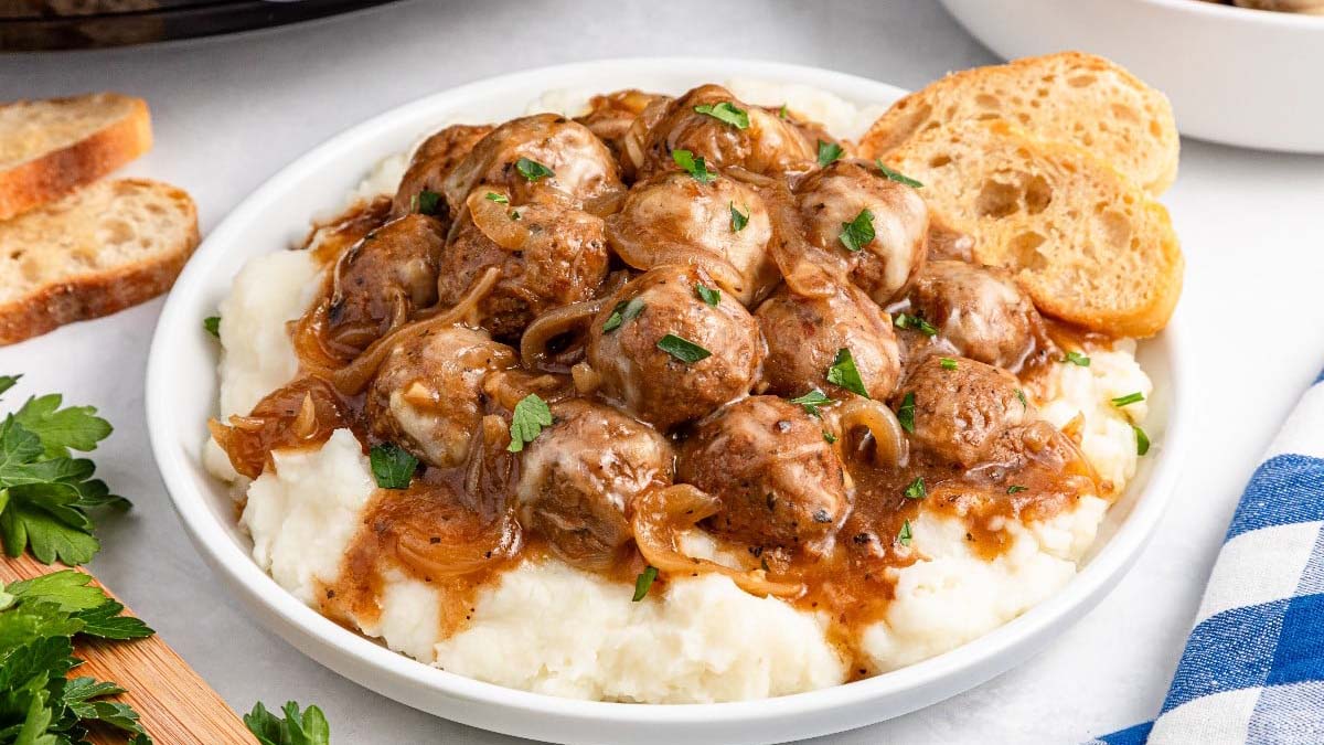 A bowl of mashed potatoes topped with meatballs, brown gravy with onions, chopped parsley, and two slices of toasted bread on the side.