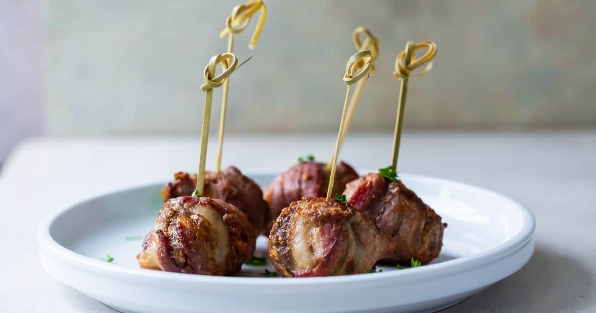 Five bacon-wrapped meatballs are served on a white plate, each held with a decorative skewer.