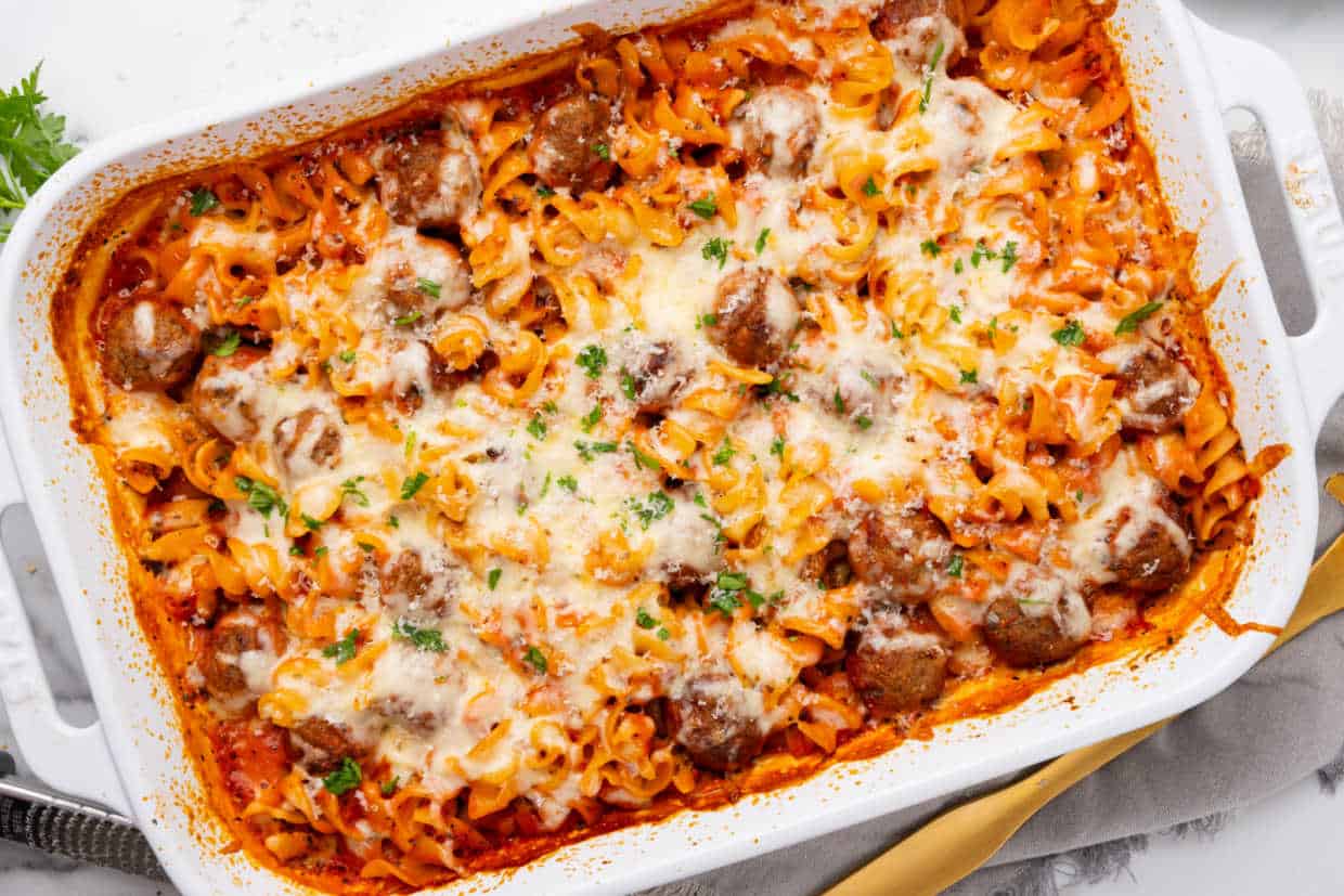 Baked pasta casserole with meatballs, tomato sauce, and melted cheese in a white baking dish, garnished with chopped herbs.