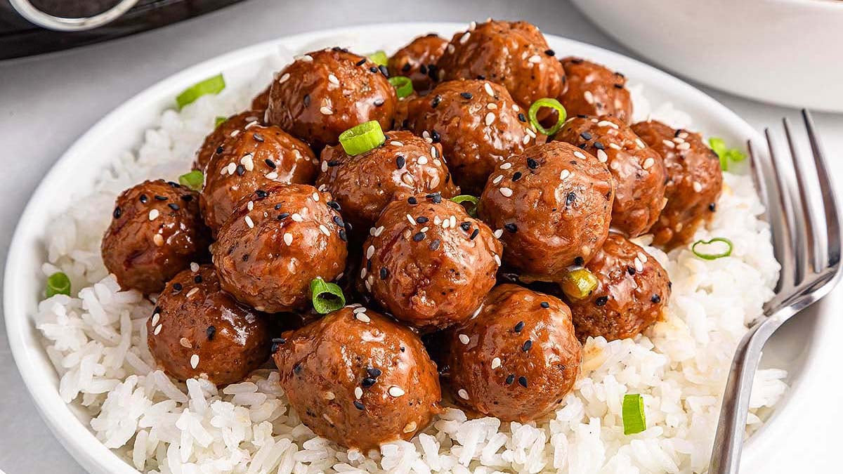 A plate of white rice topped with glazed meatballs, garnished with sliced green onions and sesame seeds, with a fork on the side.