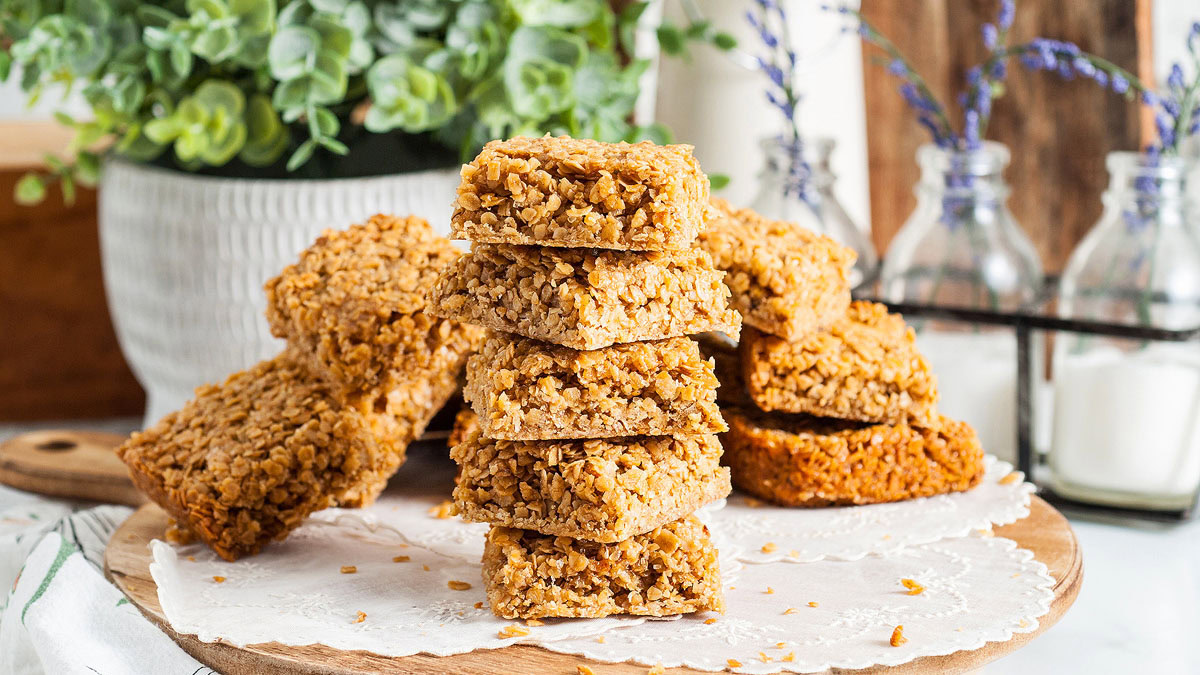Several oat flapjacks are stacked and arranged on a wooden board covered with parchment paper, with plants and bottles in the background.