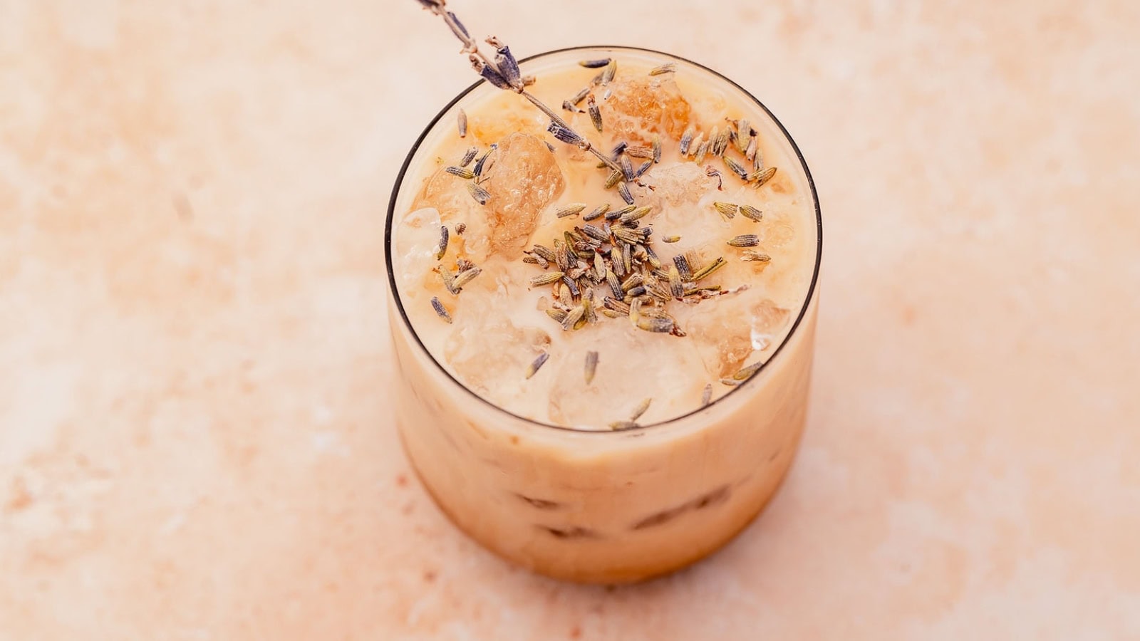 A glass of iced coffee with ice cubes, topped with dried lavender buds and a sprig of lavender on a light-colored surface.