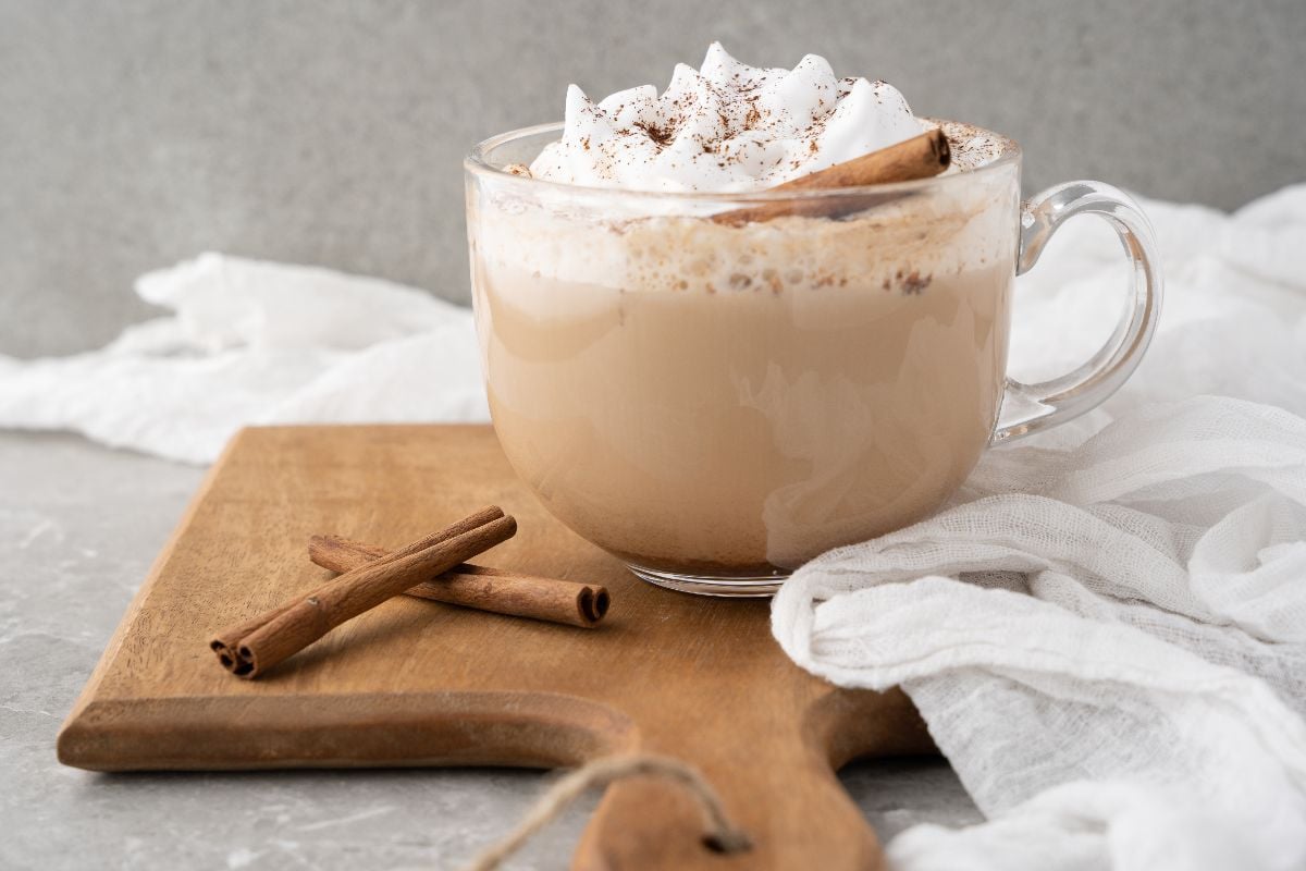 A glass cup of latte with whipped cream and a cinnamon stick sits on a wooden board, with two cinnamon sticks beside it and a white cloth nearby.