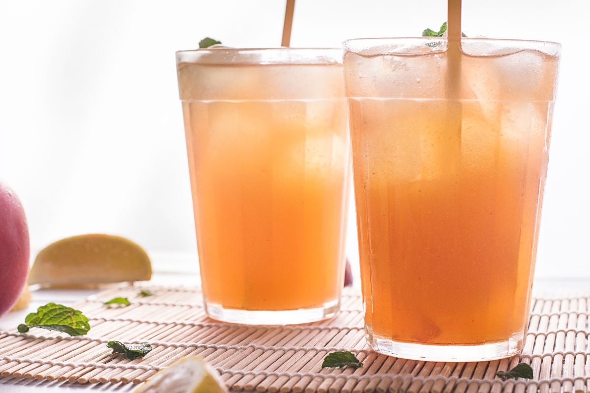 Two glasses of iced orange-colored beverage with straws and mint leaves, placed on a bamboo mat with lemon wedges and mint nearby.