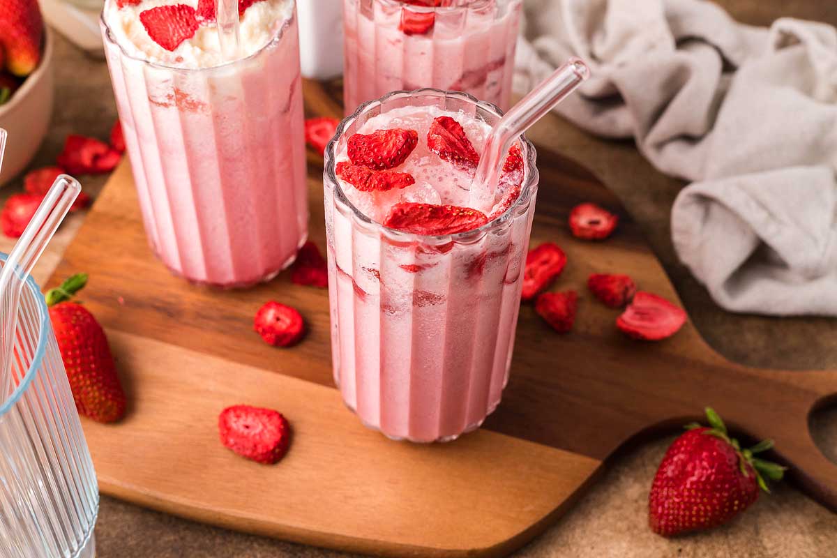 Three glasses of pink strawberry milk topped with sliced strawberries, served on a wooden board with fresh and dried strawberries scattered around.
