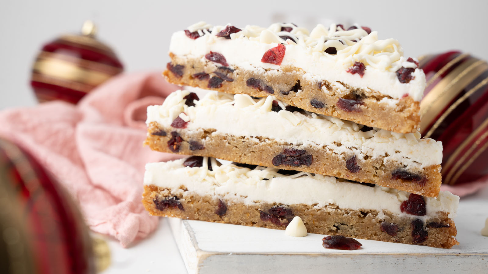 Three stacked dessert bars with a white frosting layer, white chocolate chips, and dried cranberries, displayed on a white surface with holiday ornaments in the background.
