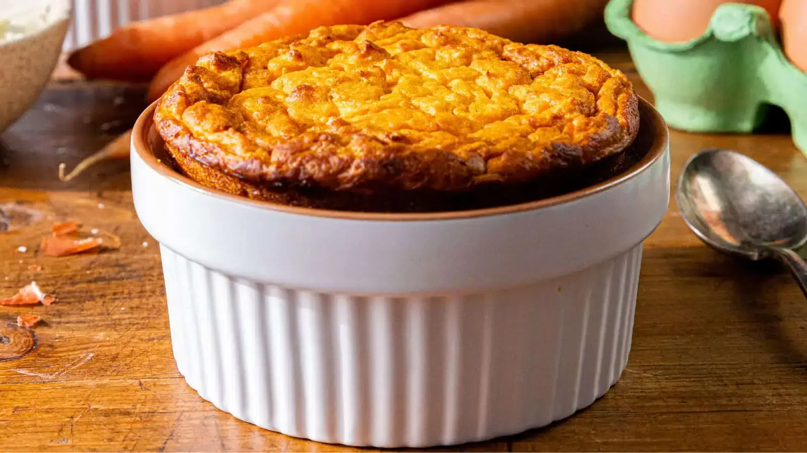 A golden-brown souffle sits in a white ramekin on a wooden surface, with eggs, carrots, and a spoon in the background.