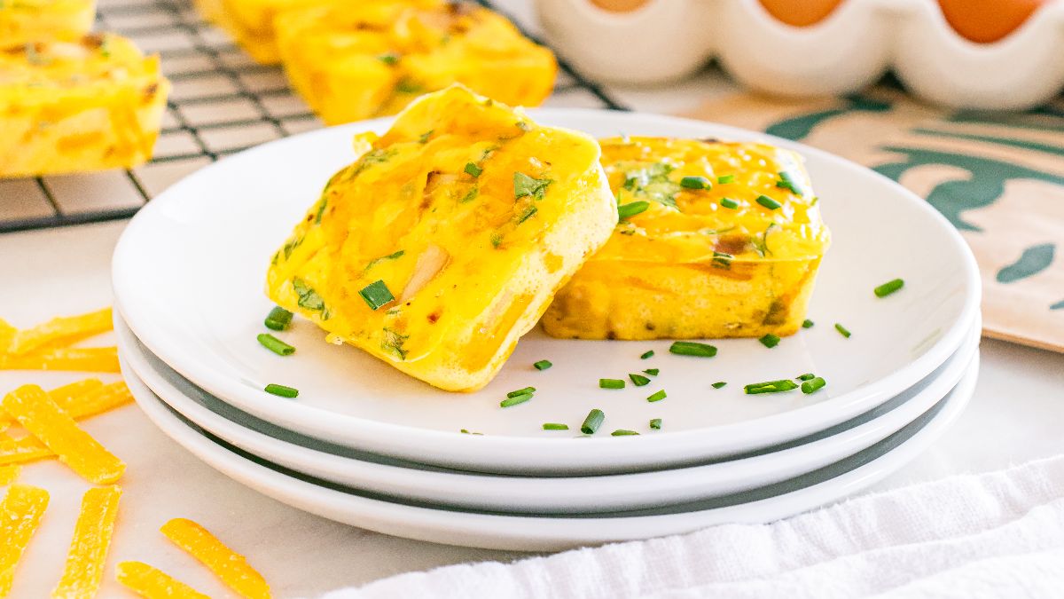 Two square egg muffins garnished with chopped chives are served on stacked white plates, with more muffins cooling in the background.