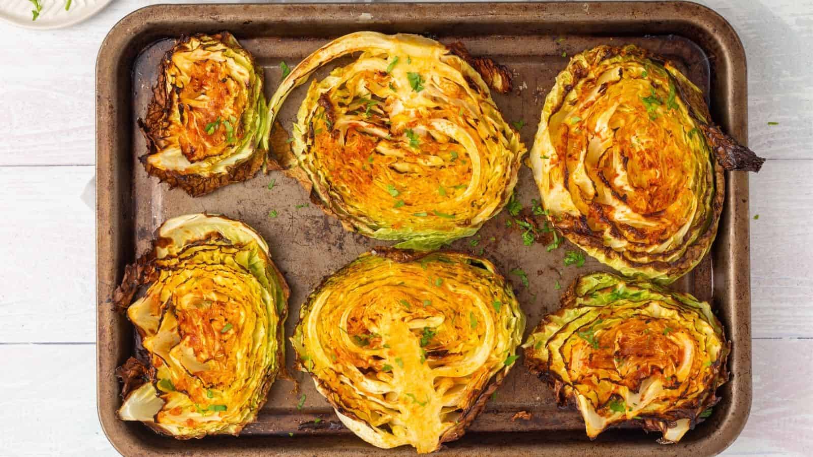 Six roasted cabbage steaks on a metal baking tray, seasoned and garnished with herbs, shown from above.