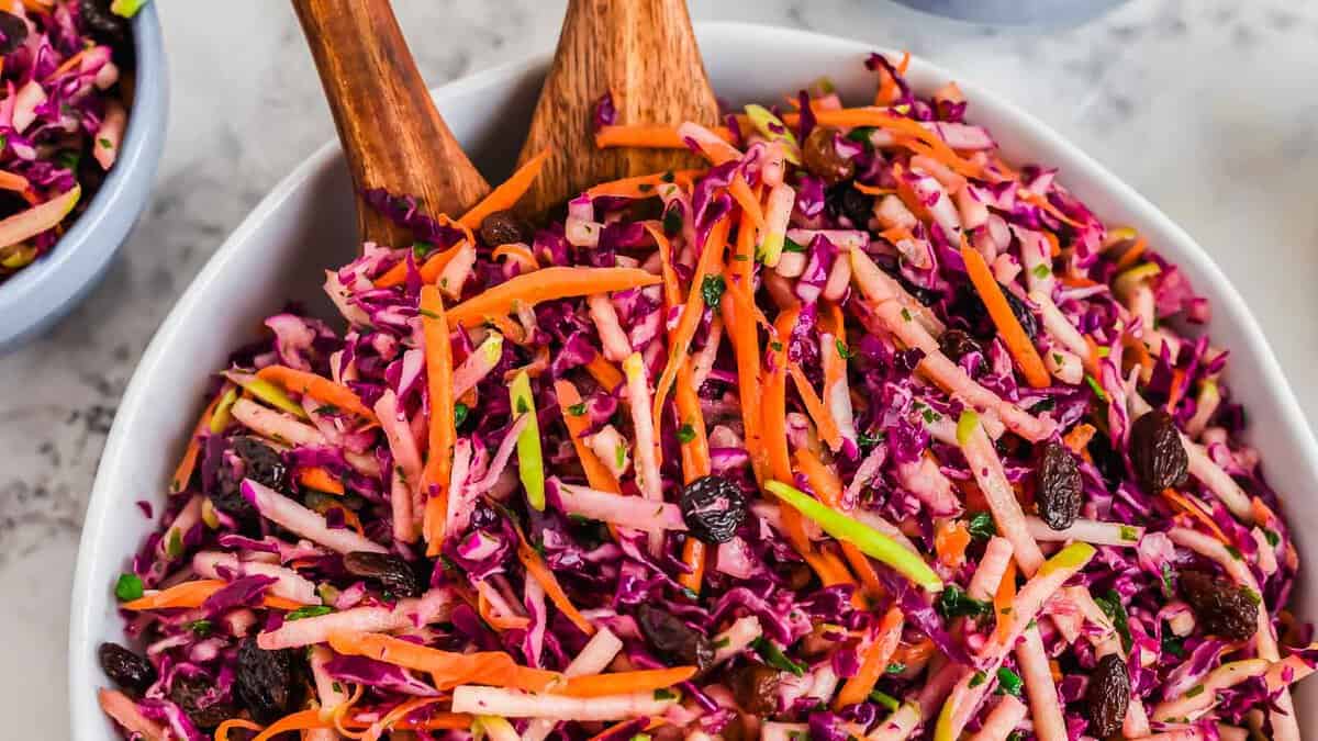 A bowl of colorful coleslaw with shredded red cabbage, carrots, green apple, and raisins, mixed with dressing and served with wooden salad tongs.