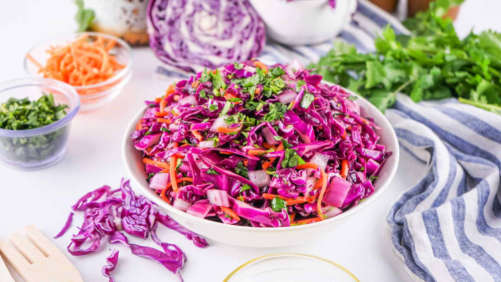 A bowl of red cabbage slaw with shredded carrots, parsley, and onions, surrounded by fresh vegetables and herbs on a white table.