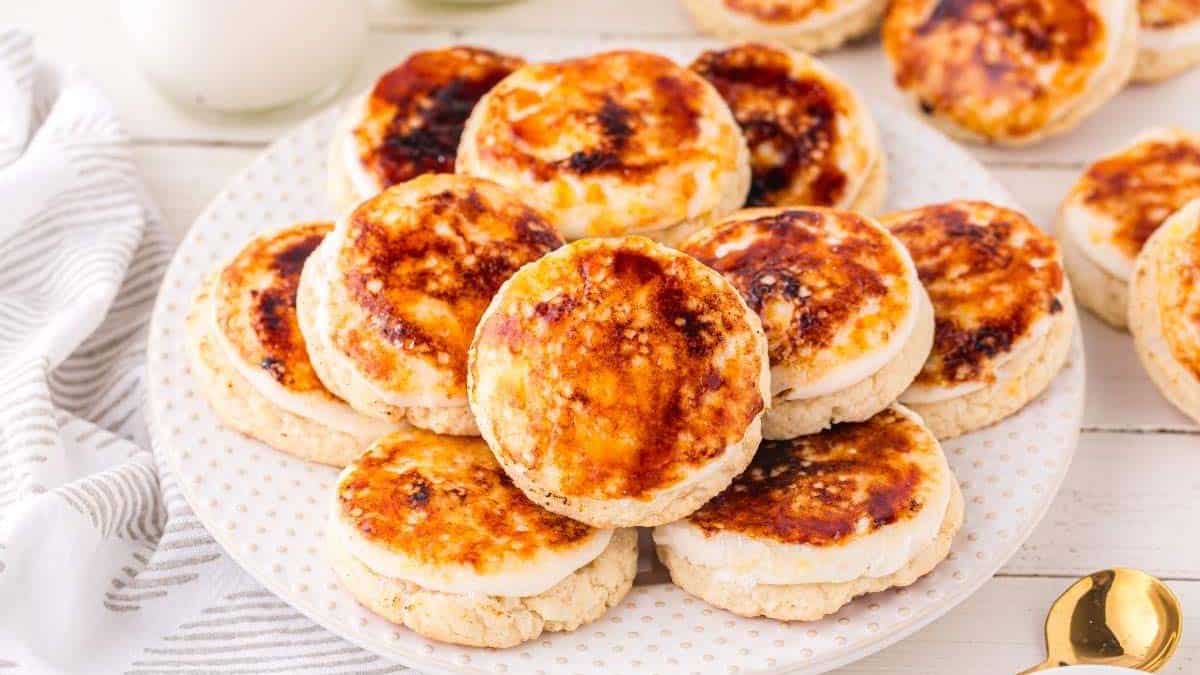 A white plate piled with round, golden-brown pastries that have caramelized tops, set on a white table with a striped cloth nearby.
