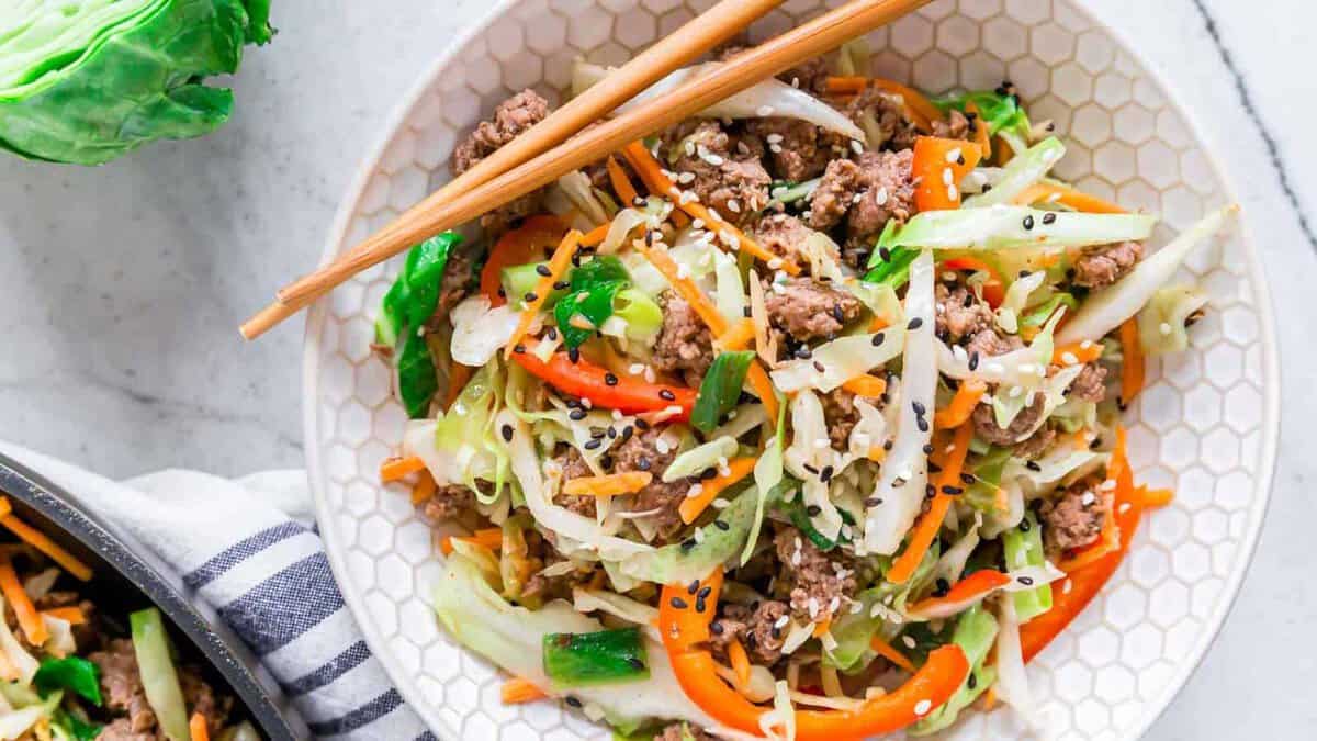 A bowl of stir-fried ground meat, cabbage, carrots, and bell peppers, topped with sesame seeds, with chopsticks resting on the rim.