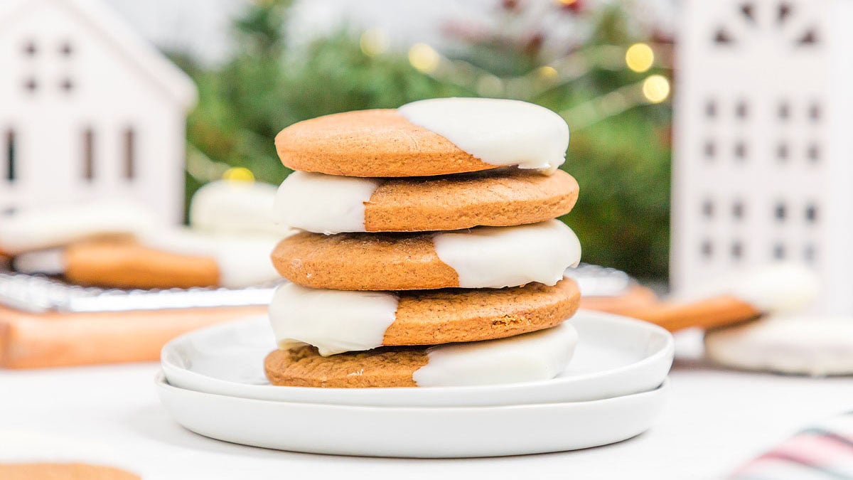 A stack of four round cookies partially dipped in white icing sits on a white plate, with blurred holiday decor in the background.