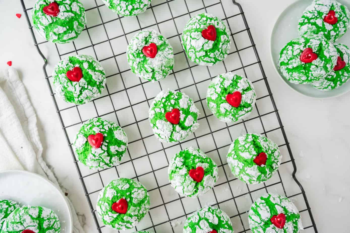 Green crinkle cookies topped with red heart candies are arranged on a black cooling rack, with several cookies on white plates nearby.