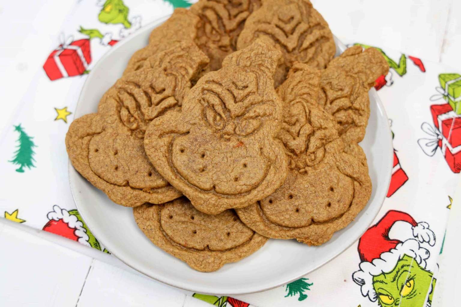 A plate of cookies shaped and decorated like the Grinch, placed on a festive napkin with holiday-themed Grinch illustrations.