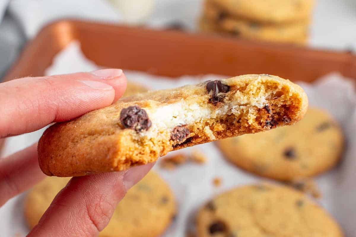 A hand holds a chocolate chip cookie with a creamy white filling, showing a bite taken out of it. Other cookies are visible on a baking tray in the background.