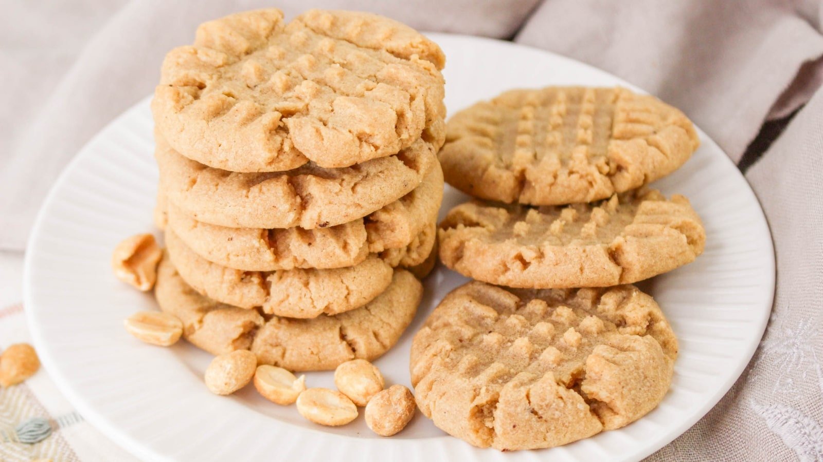 A plate of peanut butter cookies with a crisscross pattern, stacked alongside a few loose peanuts on a white plate.
