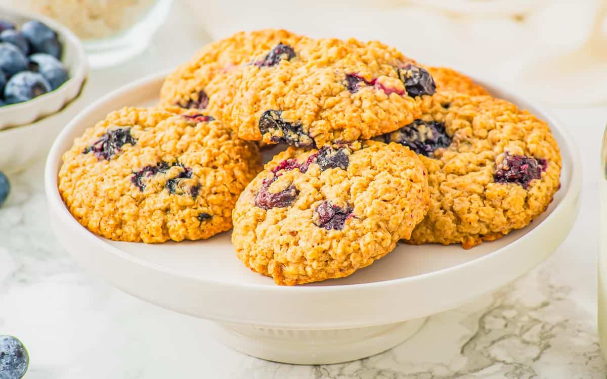 A white cake stand holds several oatmeal cookies with visible blueberries, with a bowl of fresh blueberries in the background.