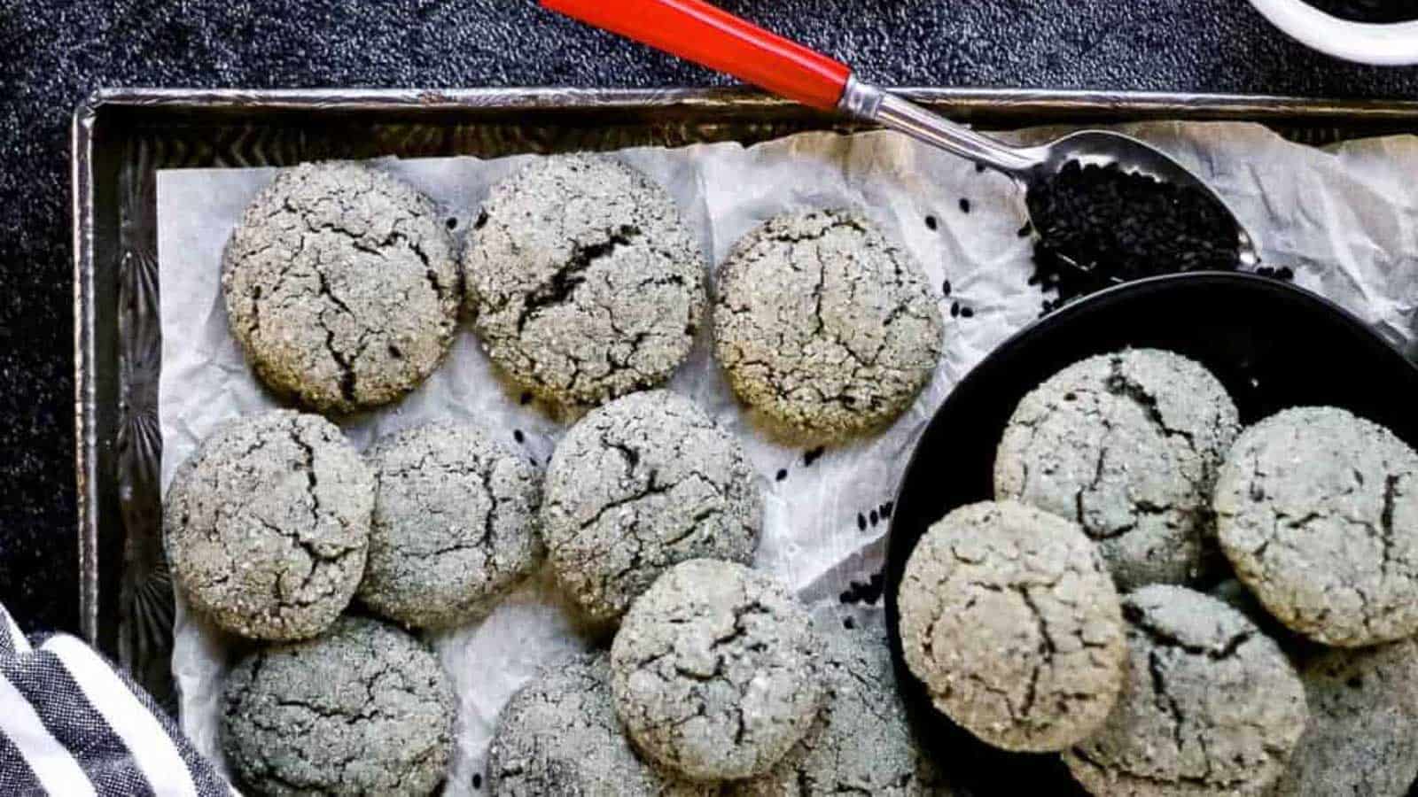 A tray of cracked, round black sesame cookies on parchment paper, with a bowl of cookies and a spoon filled with black sesame seeds beside them.