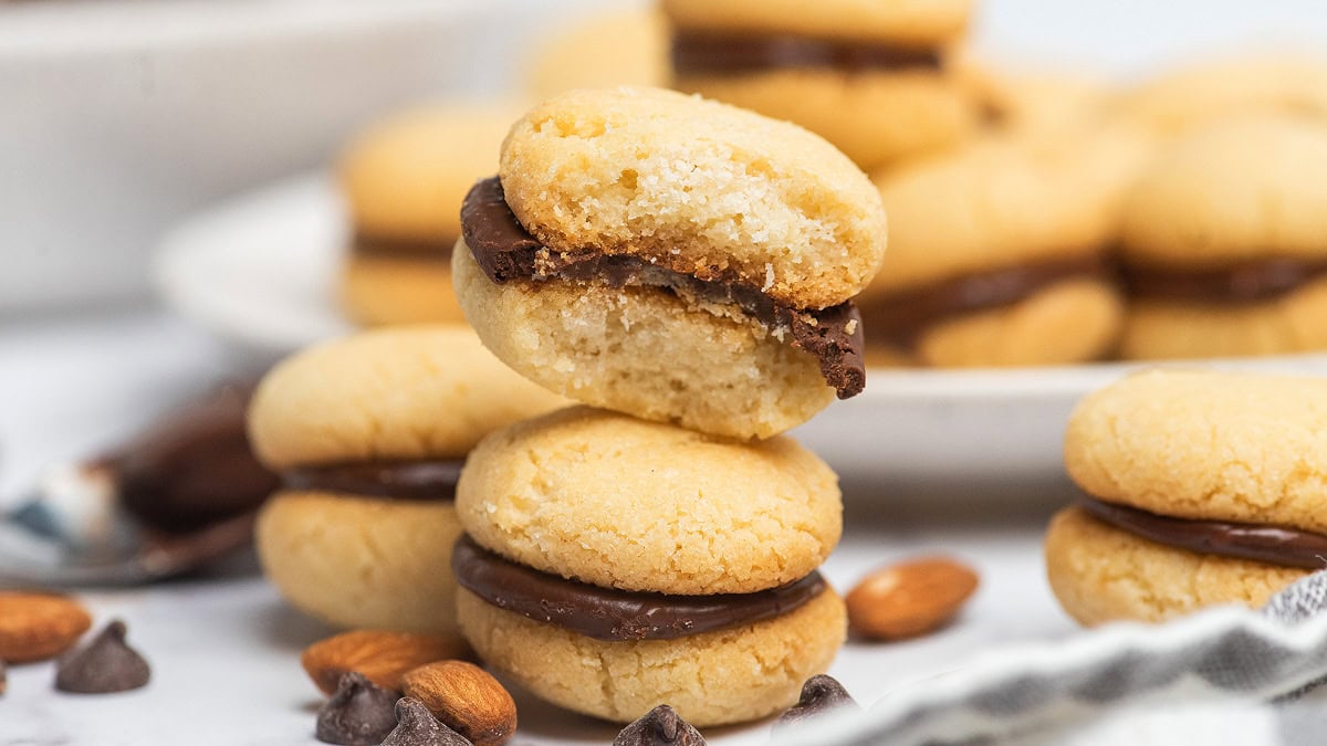 A stack of round sandwich cookies filled with chocolate cream is shown, surrounded by scattered chocolate chips and almonds on a white surface.