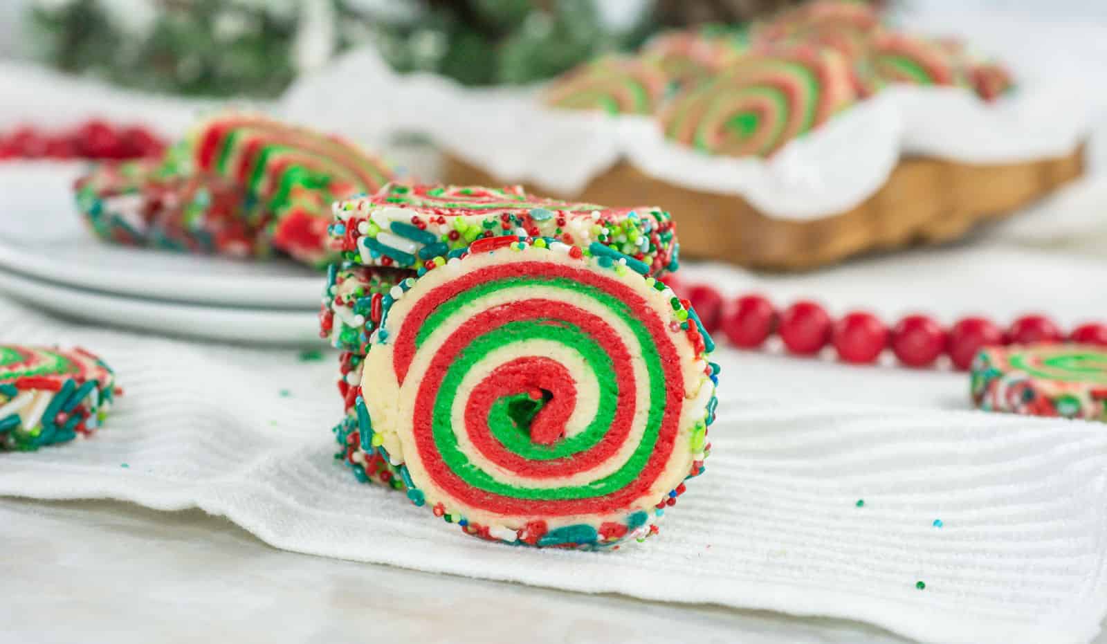 A close-up of a spiral sugar cookie with red, green, and white swirls, coated with colorful sprinkles, placed on a white cloth with more cookies in the background.