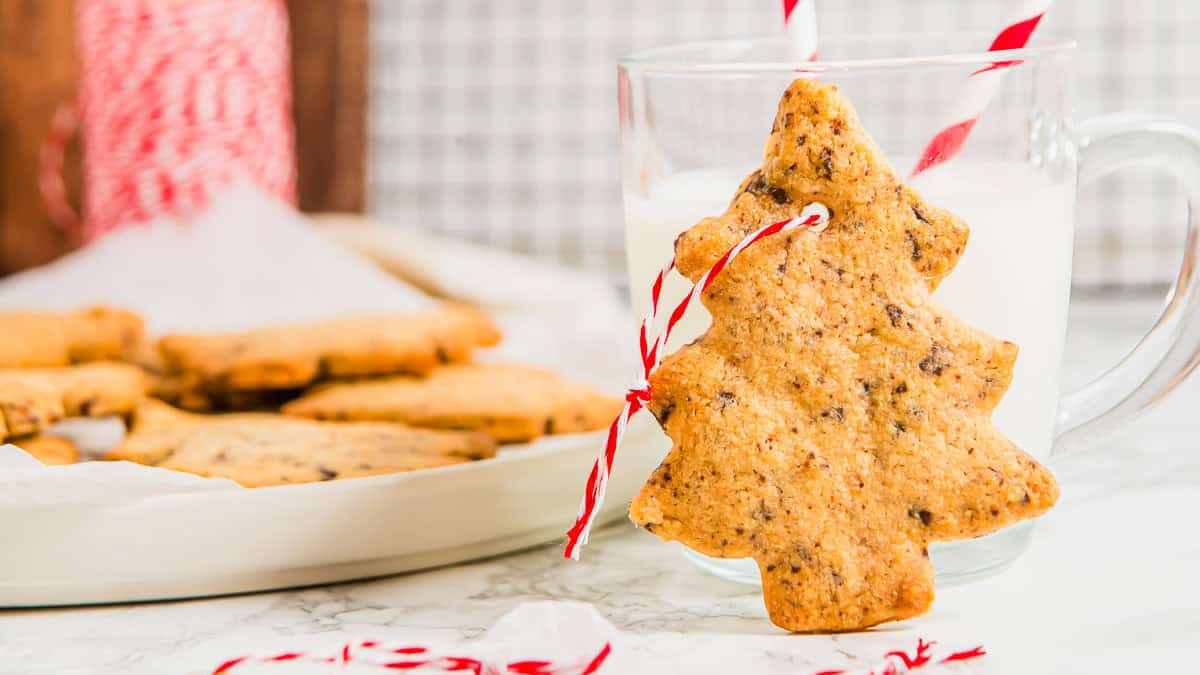 A tree-shaped cookie with a red and white string hangs on a glass of milk; more cookies are on a plate in the background.