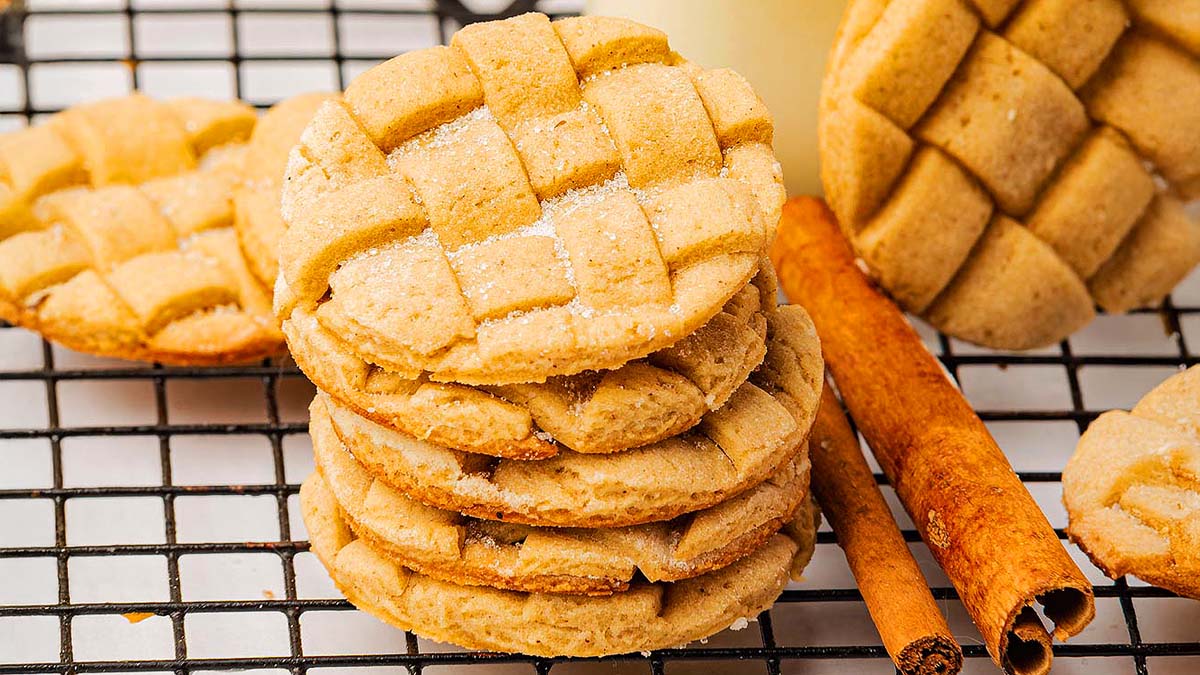 A stack of lattice-patterned cookies sits on a cooling rack next to two cinnamon sticks.