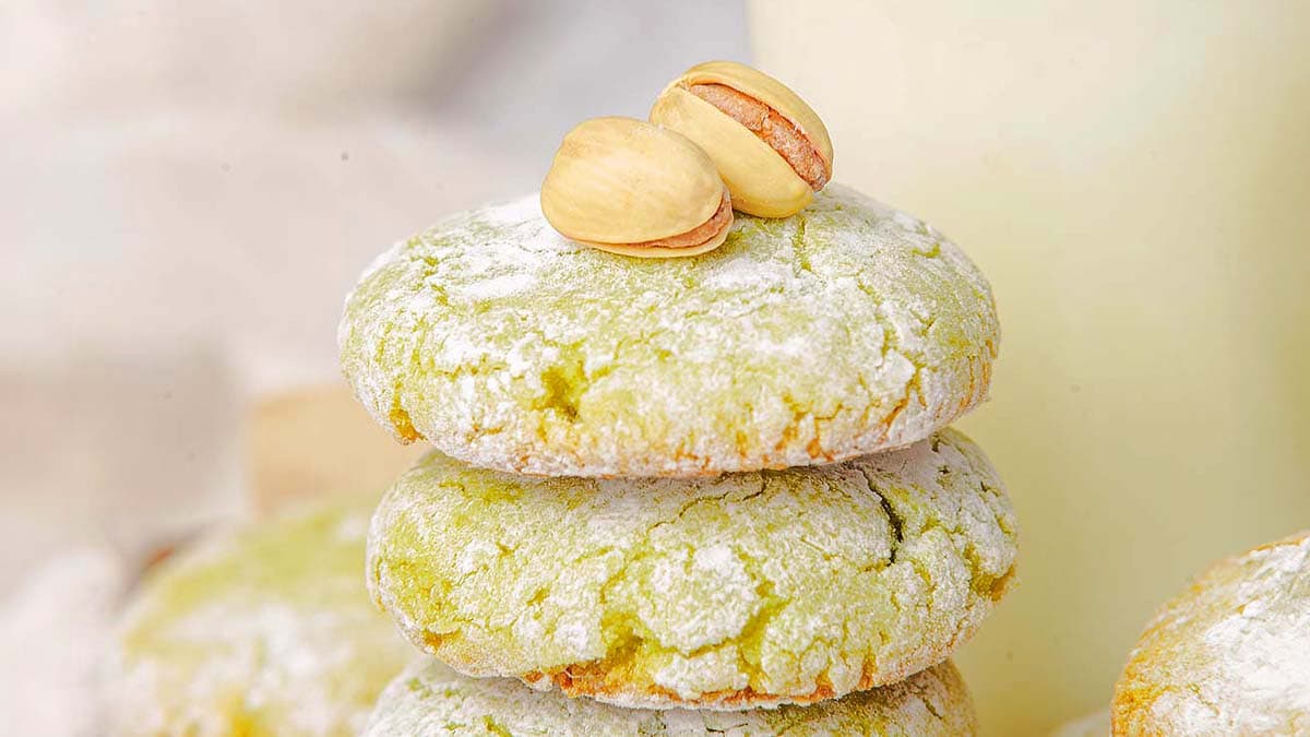 Three stacked pistachio cookies dusted with powdered sugar, topped with two whole pistachio nuts, with more cookies in the blurred background.