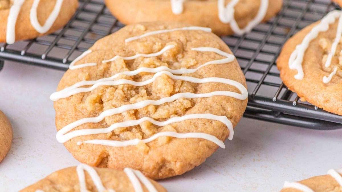 Close-up of round cookies with crumb topping and white icing drizzle, cooling on a black wire rack.