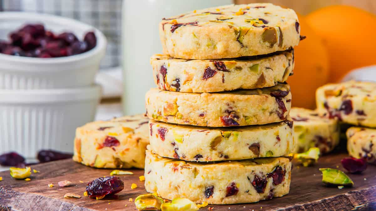 A stack of cranberry pistachio cookies on a wooden board, with chopped pistachios and cranberries scattered around.