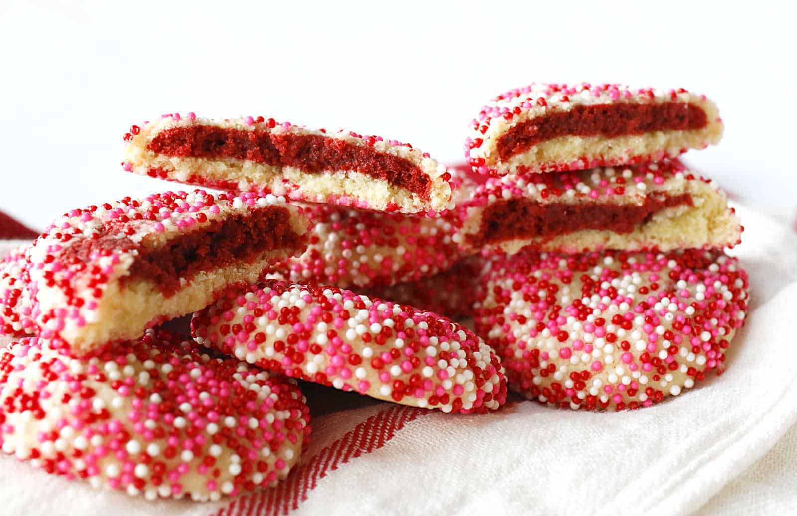 Sugar cookies covered in pink, red, and white sprinkles are stacked on a cloth; some are halved to reveal a red filling inside.