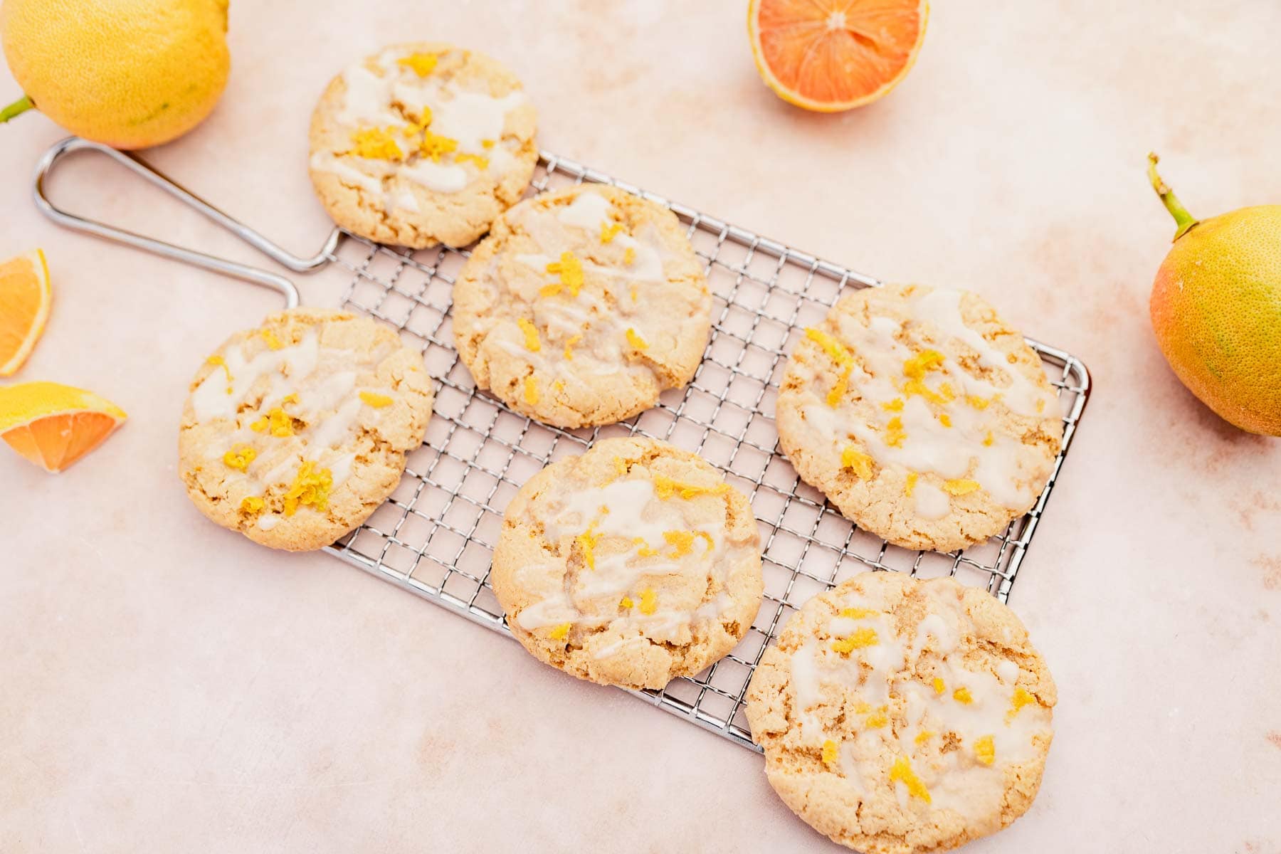 Six iced cookies with orange zest sit on a cooling rack, surrounded by whole and sliced oranges on a light surface.