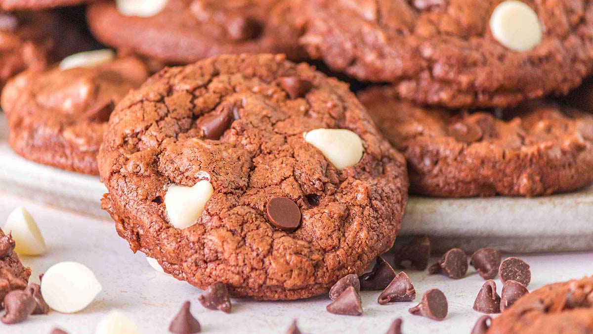 A close-up of chocolate cookies with white and milk chocolate chips on a plate, with extra chocolate chips scattered nearby.