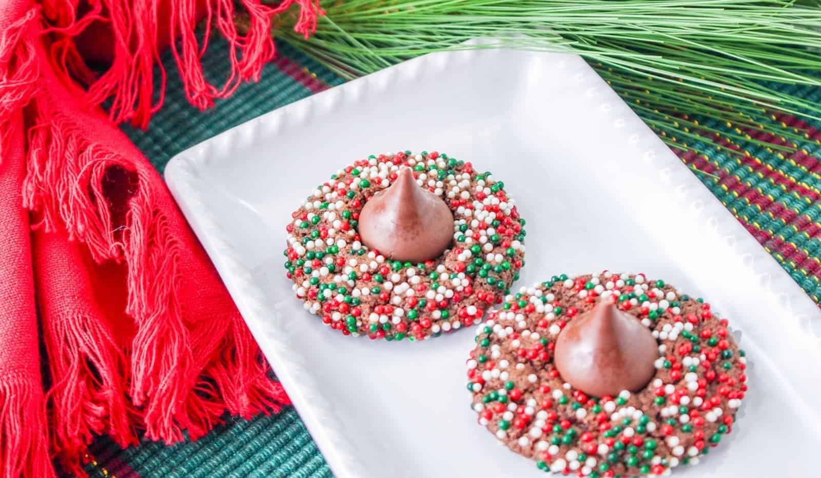 Two chocolate kiss cookies covered in red, white, and green sprinkles on a white rectangular plate, with red fabric and greenery in the background.
