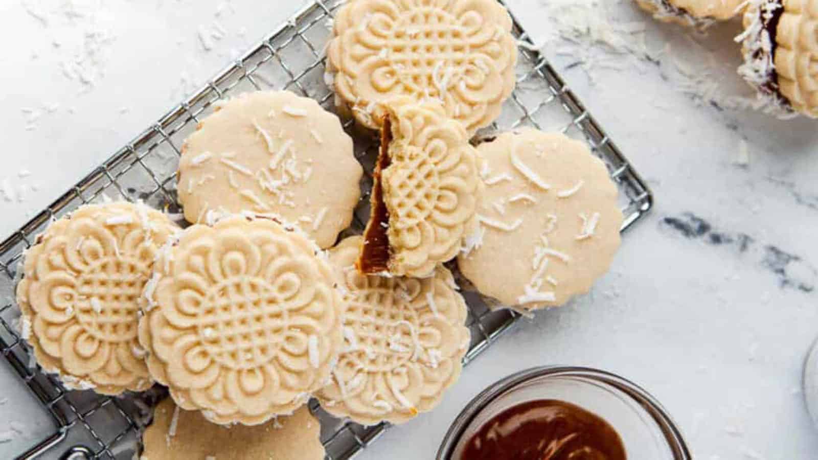 A stack of floral-patterned sandwich cookies with a caramel filling sits on a wire rack, sprinkled with shredded coconut. A small bowl of caramel is nearby.