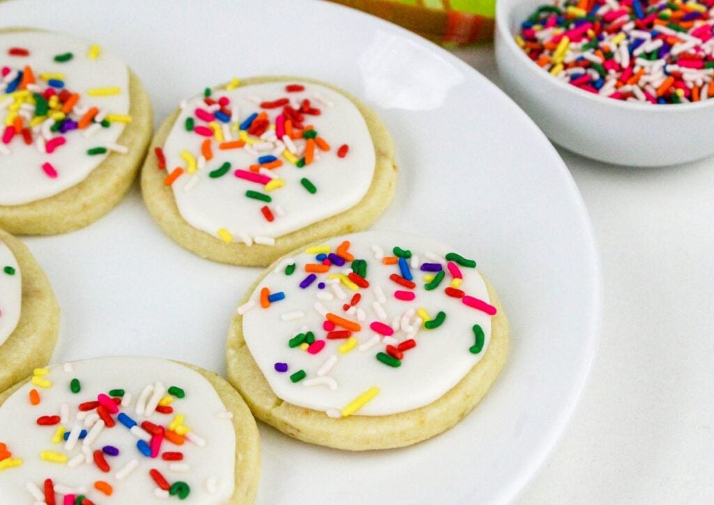 Sugar cookies with white icing and colorful sprinkles are arranged on a white plate, with a bowl of extra sprinkles nearby.