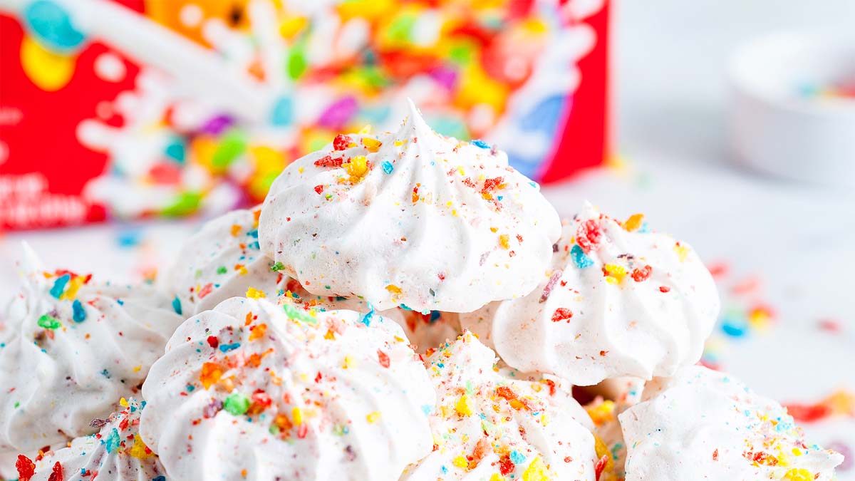 A pile of white meringue cookies topped with colorful cereal crumbs, with a blurred box of cereal in the background.