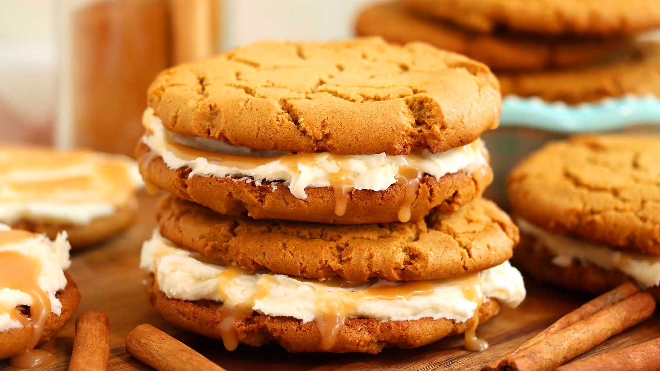 Two cookie sandwiches with cream filling and caramel sauce are stacked on a wooden board, with cinnamon sticks nearby and more cookies in the background.