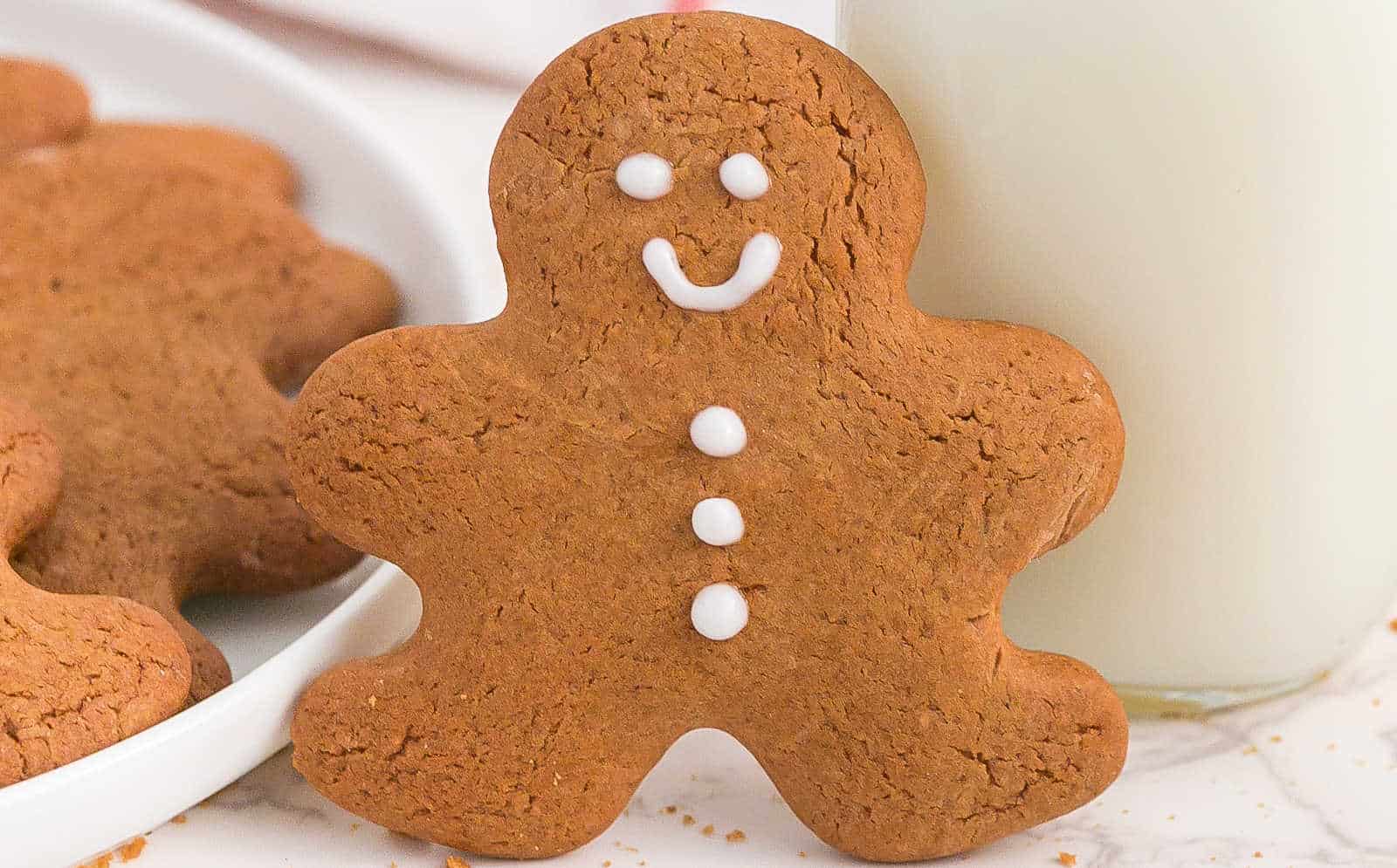 A gingerbread cookie shaped like a person, decorated with white icing for the face and buttons, stands next to a glass of milk.