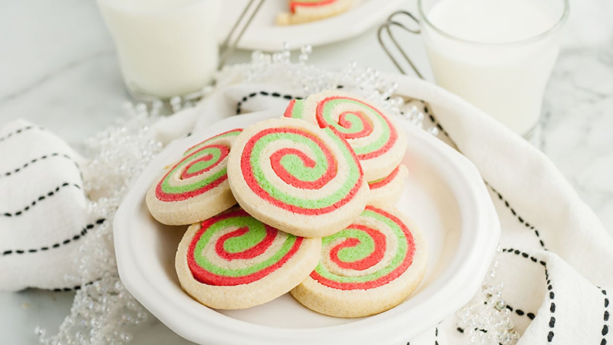 A plate of round sugar cookies with red and green spiral patterns, placed on a white surface with glasses of milk and a white cloth nearby.
