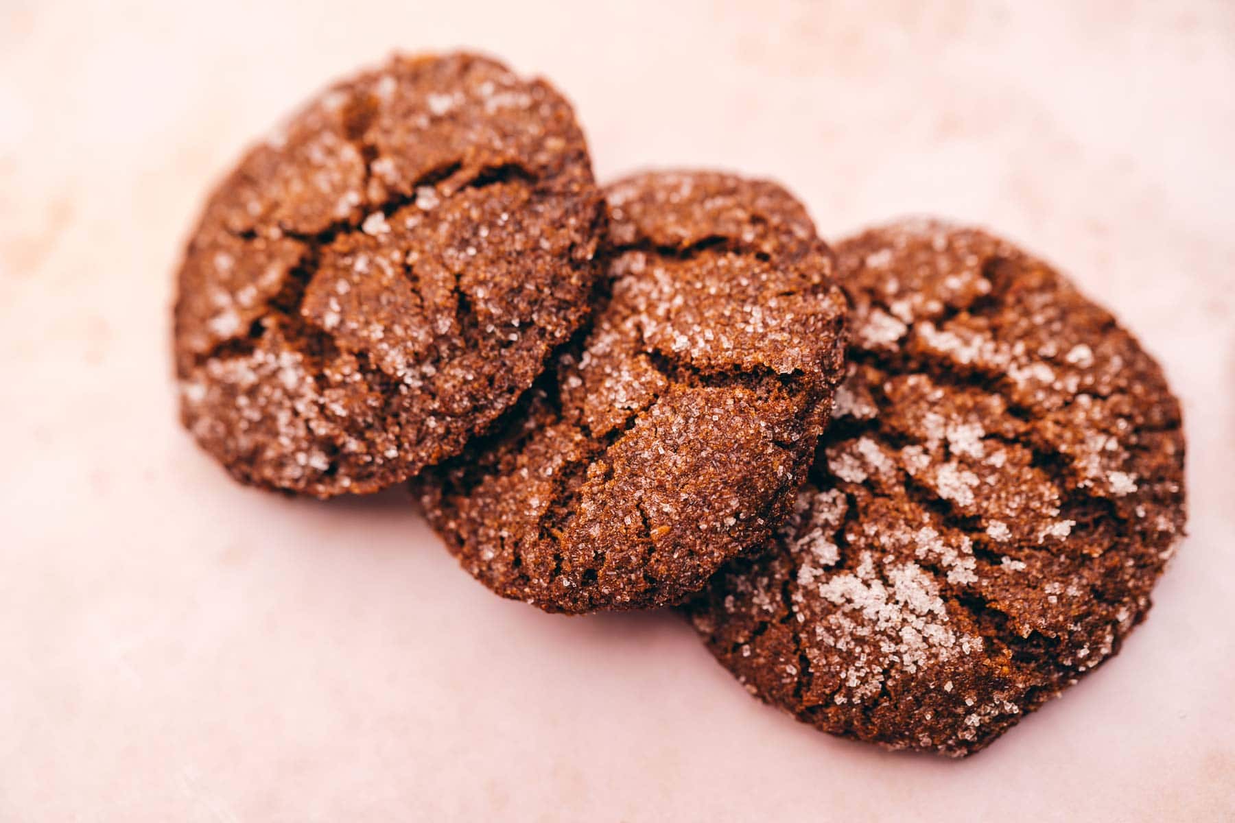 Three chocolate cookies with a cracked surface and a dusting of sugar are arranged in a row on a light-colored background.
