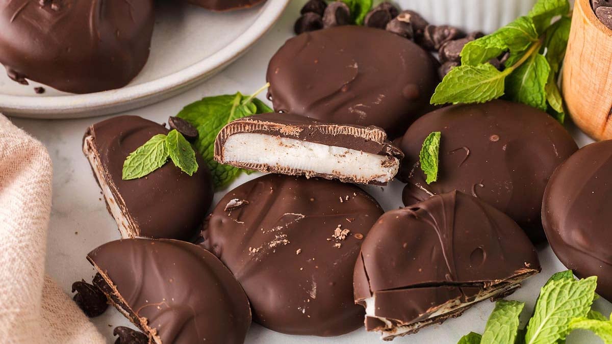 Chocolate-covered peppermint patties, some whole and some cut in half to show the white mint filling, are arranged on a surface with fresh mint leaves and chocolate chips.