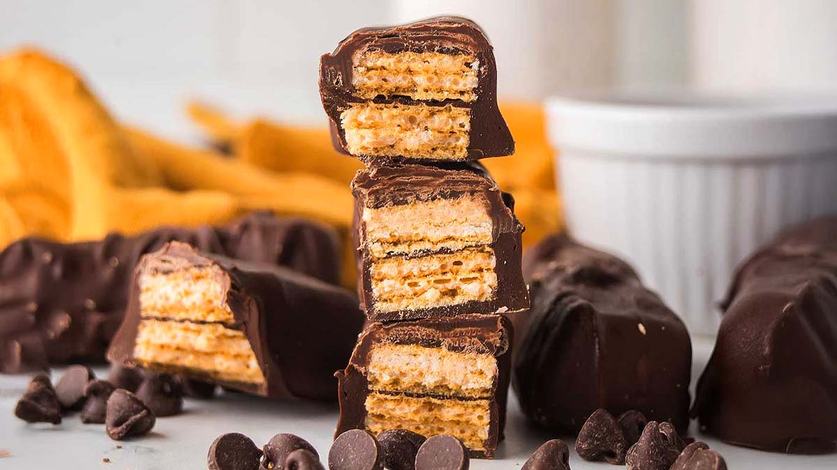 A stack of chocolate-covered wafer bars is shown with chocolate chips scattered around and a white ramekin in the background.