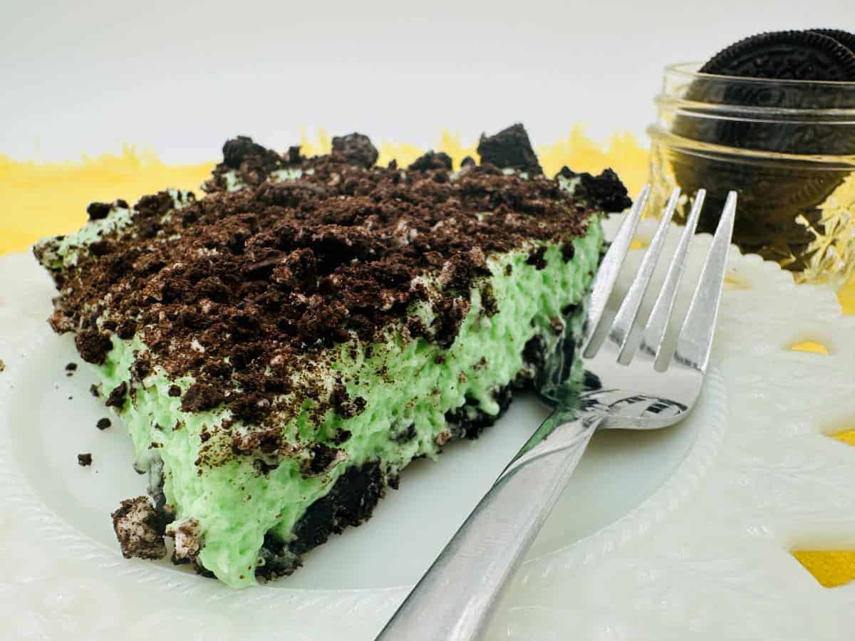 A slice of mint Oreo pie topped with crushed Oreos sits on a white plate with a fork; a jar of whole Oreo cookies is in the background.