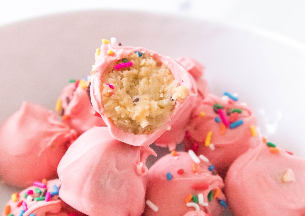 A close-up of pink, sprinkle-covered cake balls, with one showing a bitten cross-section revealing the moist cake interior.