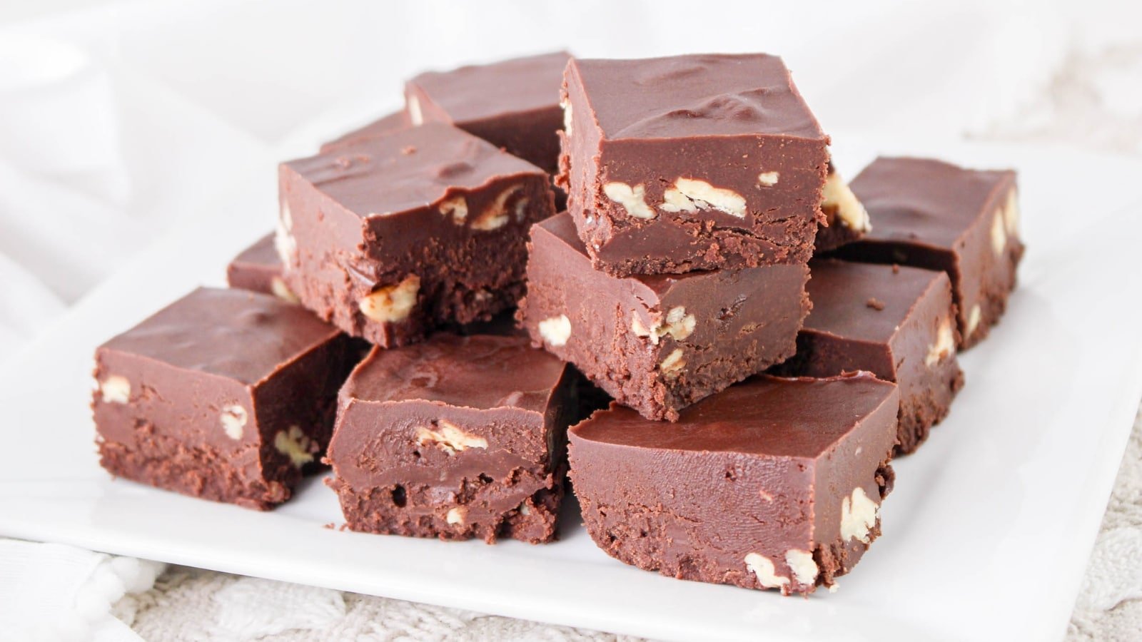 A plate of chocolate fudge squares with visible pieces of nuts, stacked on a white dish.