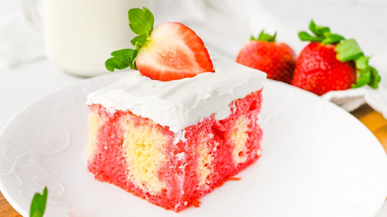 A slice of strawberry poke cake with white frosting and a halved strawberry on top, served on a white plate with whole strawberries in the background.