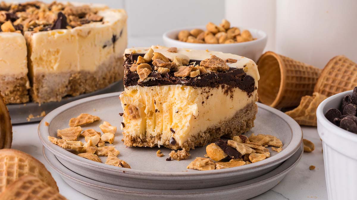 A slice of ice cream cake with a chocolate topping and chopped peanuts sits on a plate, surrounded by waffle cones, peanuts, and a bowl of chocolate chips.